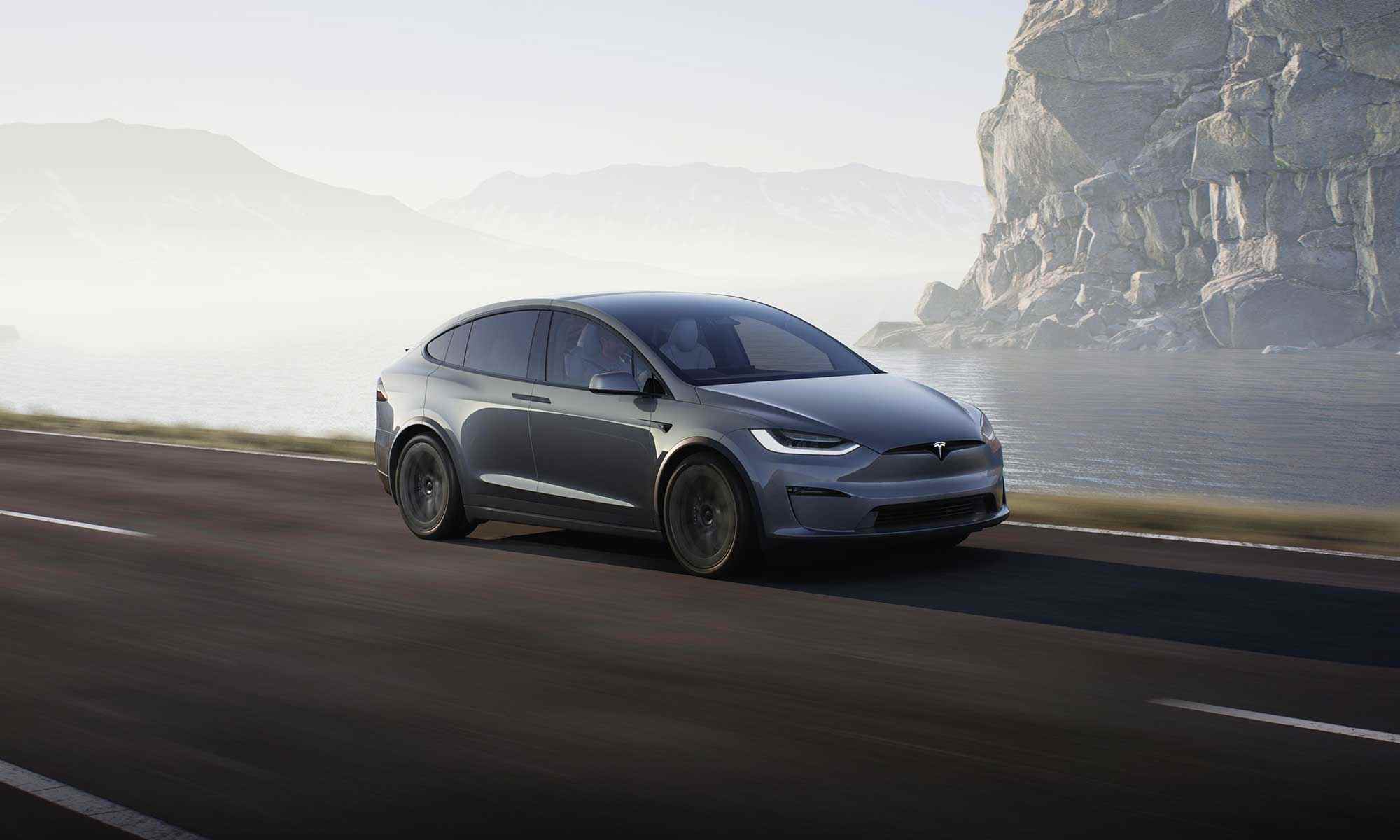 dark grey Tesla model X driving down a country road, past a lake and mountains