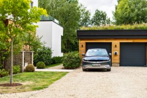 Wide shot of a gravel driveway, a double garage, with a Pod Point Solo installed in-between the two doors, and plugged in to a dark blue EV.