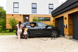 A wide shot of a mother and two daughters plugging the cable into their black EV, which is parked in front of the garage doors, where the Pod Point Solo 3 is installed.