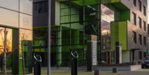 Wide shot of two Pod Point twin chargers in front of a modern glass commercial building at sunset.