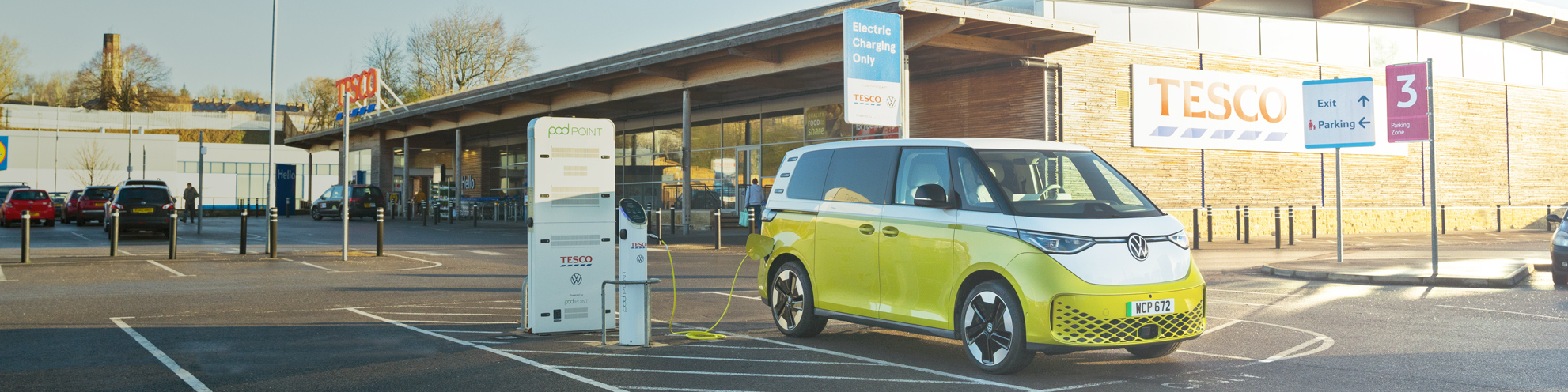Wide shot of a yellow VW camper van plugged in to a Pod Point twin charger in front of Tesco
