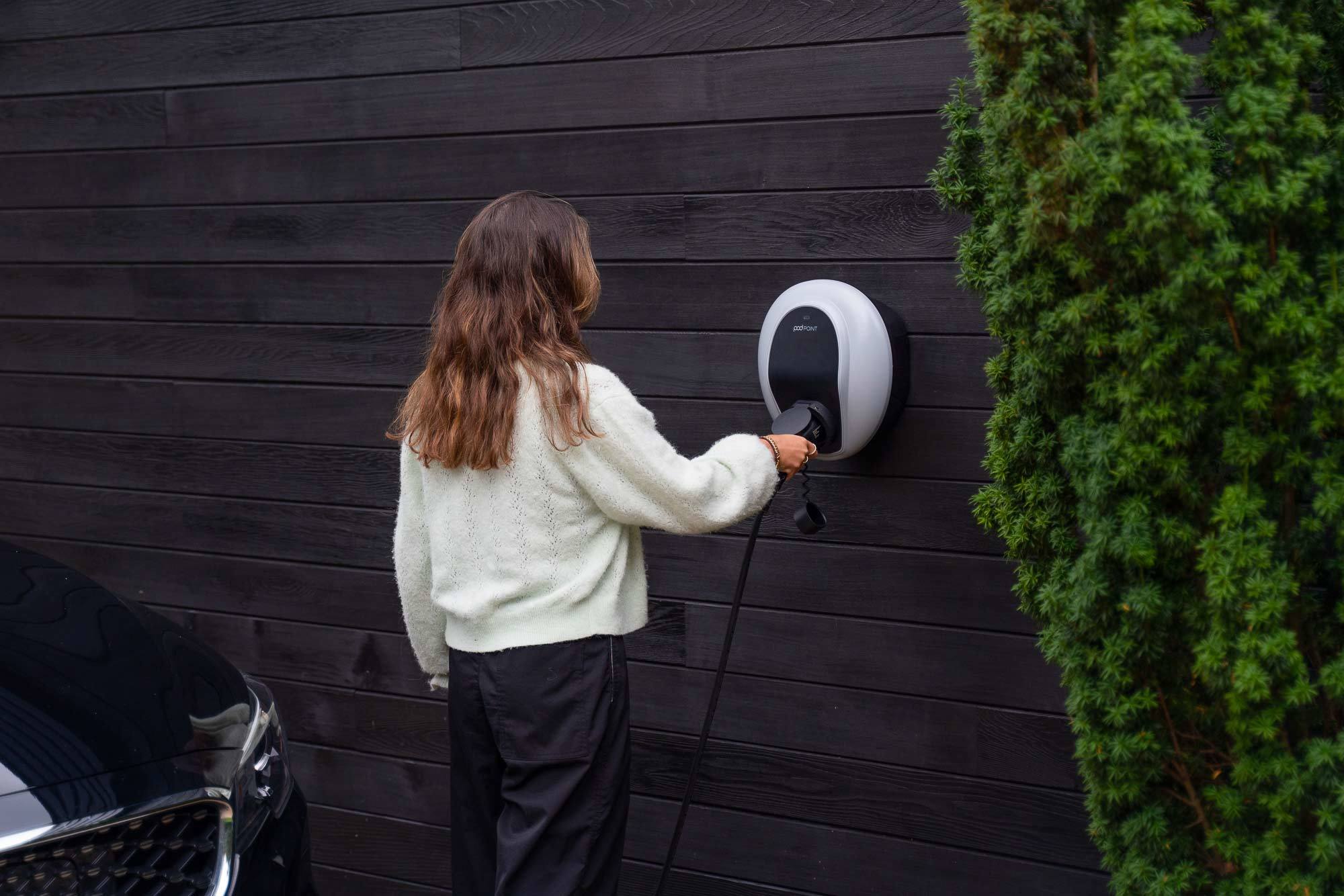 A Solo 3 installed on a black wooden wall, with a tall hedge to the right. A woman with mid-length brown hair and a white jumper is plugging a cable in to the charger, with her back to us