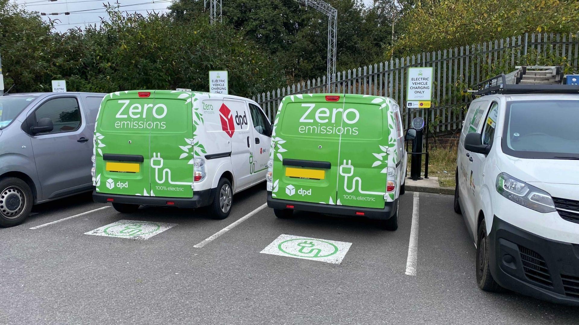 Two DPD electric vans with bright green back doors and 'zero emissions' written on the back, parked in EV charging bays in a busy car park
