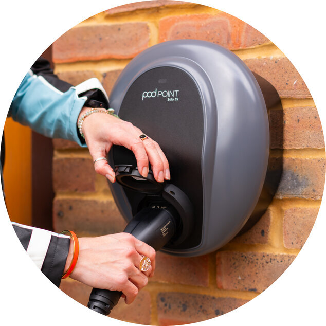 A Pod Point Solo 3 installed on a dark wood wall, with an unfocused hand checking their phone in the forefront